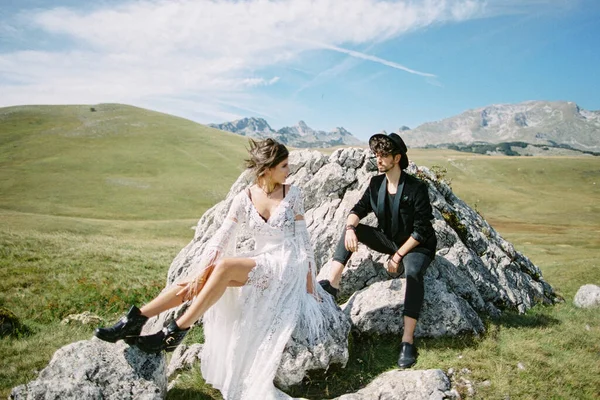 Novia y novio están sentados sobre piedras en un césped verde contra el telón de fondo de las montañas — Foto de Stock