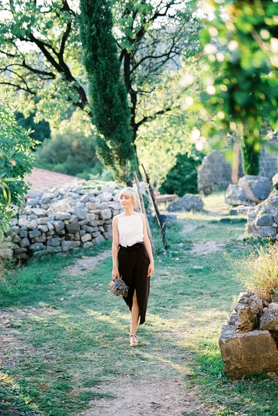 Mulher com um buquê de lavanda caminha ao longo de um caminho em um parque verde — Fotografia de Stock