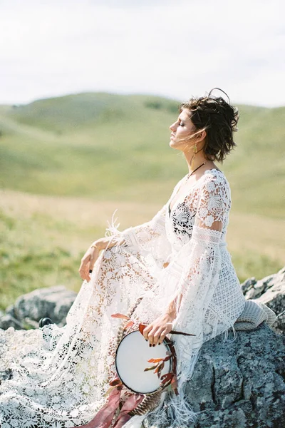 Fille dans une robe blanche avec un tambourin dans sa main est assis sur des pierres sur une pelouse verte — Photo