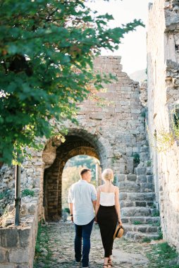 Bir adam ve bir kadın kalenin taş duvarının yanında el ele yürüyorlar. Arka plan