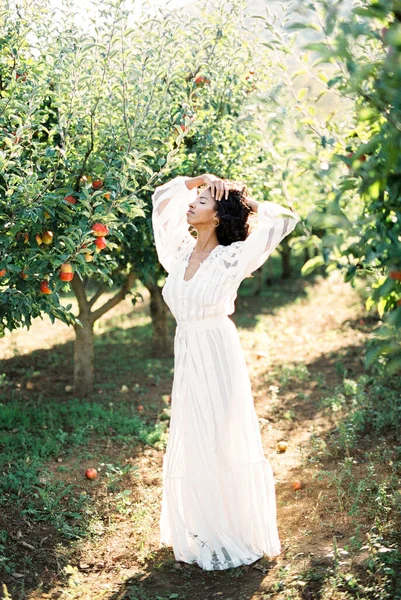 白いドレスの女性は彼女の頭の後ろに彼女の手でリンゴの果樹園に立っている — ストック写真