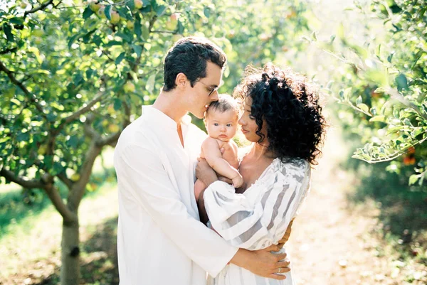 Homem abraçando uma mulher em um vestido branco com um bebê em seus braços no jardim Fotos De Bancos De Imagens