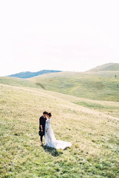 Gelin ve damat yeşil bir çimenlikte, dağların arkasında duruyorlar.
