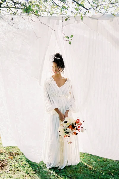 Žena v dlouhých šatech s kyticí stojí v blízkosti krajkové bílé opony v zahradě — Stock fotografie