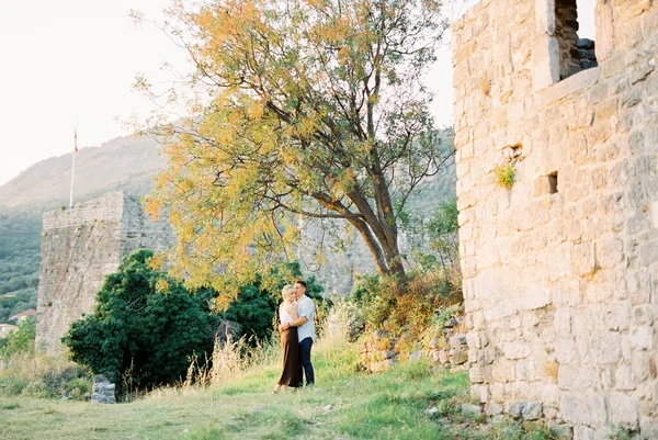 Ο άντρας αγκαλιάζει τη γυναίκα ενώ στέκεται δίπλα σε ένα δέντρο σε ένα φόντο πέτρινου τοίχου στο πάρκο. — Φωτογραφία Αρχείου