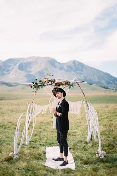 Panna młoda w czarnym kapeluszu stoi na białym dywanie przed ślubnym łukiem na trawniku — Zdjęcie stockowe