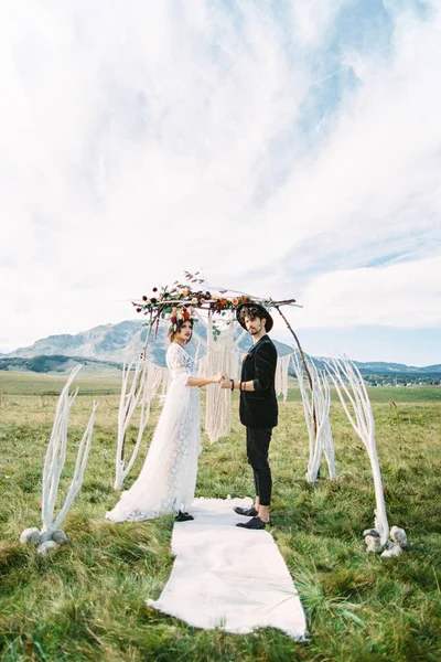 Noiva e noivo ficar em um tapete branco de mãos dadas perto do arco de casamento — Fotografia de Stock