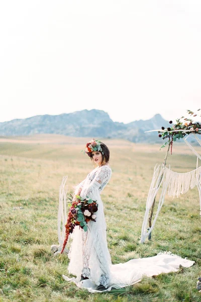 La sposa in una ghirlanda con un mazzo sta su un tappeto bianco vicino a un arco nuziale — Foto Stock
