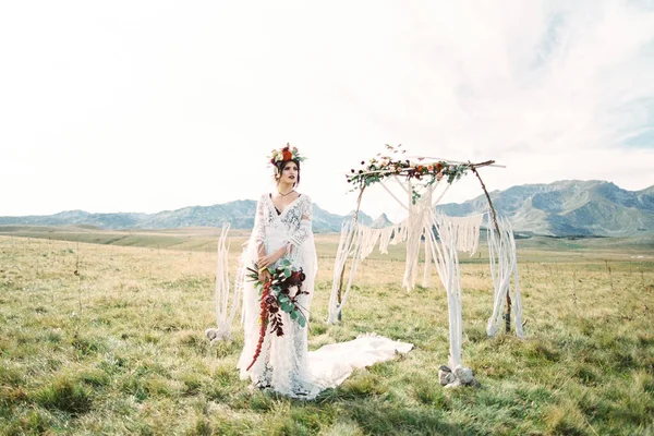 Pengantin dalam karangan bunga dengan buket berdiri di dekat lengkungan pernikahan di halaman — Stok Foto