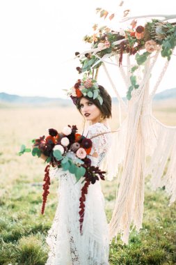 Ellerinde bir buket çiçekle gelin, düğün kemerinin yanında duruyor.