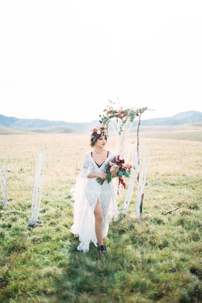 Sposa in una ghirlanda cammina oltre un arco di matrimonio su un prato verde — Foto Stock