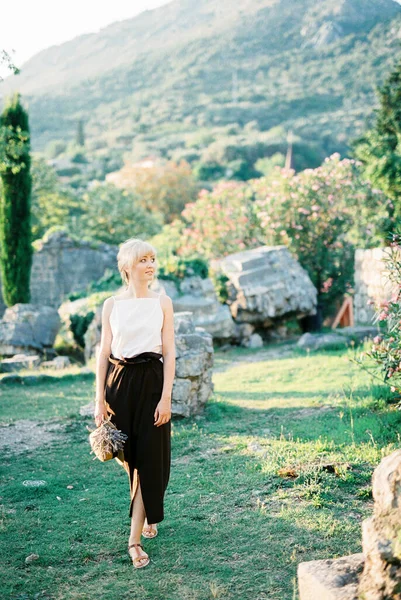 ラベンダーの花束を持つ女性は 石の遺跡を背景に緑の庭を歩いています 高品質の写真 — ストック写真