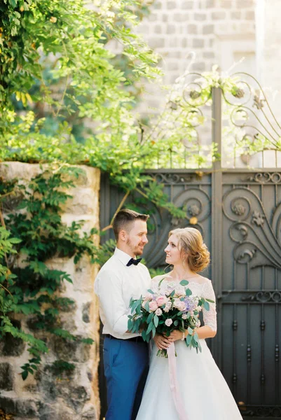 Mariée et marié se tiennent près de la porte forgée dans une clôture en pierre — Photo