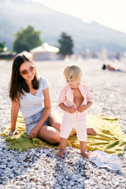 Little girl unzips her overalls while standing on the beach. High quality photo