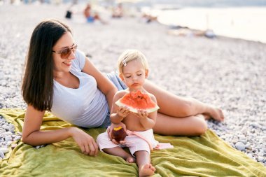Smiling mom holding out peach to little girl eating watermelon on beach. High quality photo