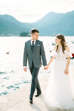Bride and groom holding hands walk along the pier against the backdrop of the mountains. High quality photo