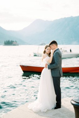 Groom hugs bride on the pier against the backdrop of boats and mountains. High quality photo