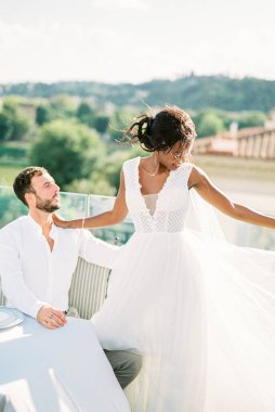 Bride stands next to sitting groom near the table on the terrace of the building. High quality photo