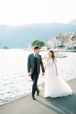 Bride and groom walk along the Perast embankment against the backdrop of the mountains. High quality photo