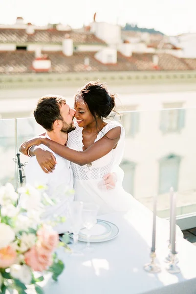 Bride sits in the arms of groom at the table on the terrace of the building. High quality photo