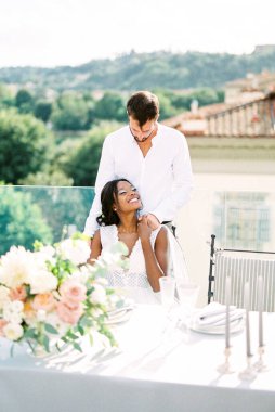 Groom hugs from behind bride sitting at the table on the terrace of the building. High quality photo