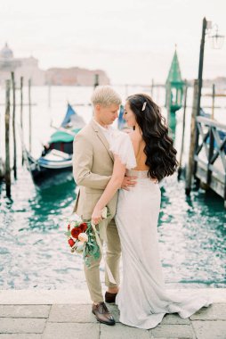 Groom hugs bride with a bouquet on the pier with gondolas in Venice. High quality photo