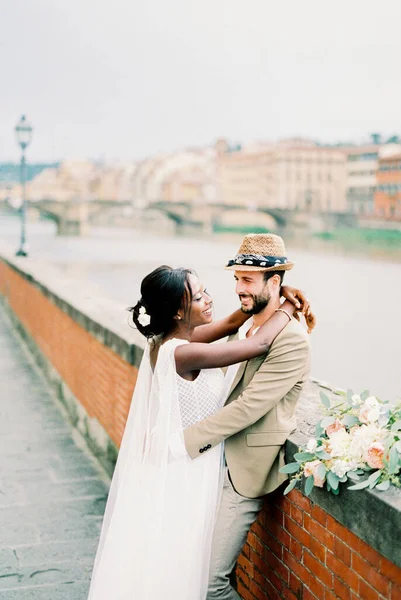 Groom Hugs Bride Stone Fence River Backdrop Buildings High Quality — Stockfoto