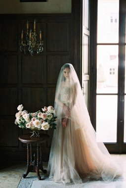 Bride in a veil stands near the table with a bouquet of flowers in a vase. High quality photo
