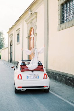Bride sits on the convertible top of a Fiat 500 car parked outside the building. Back view. High quality photo