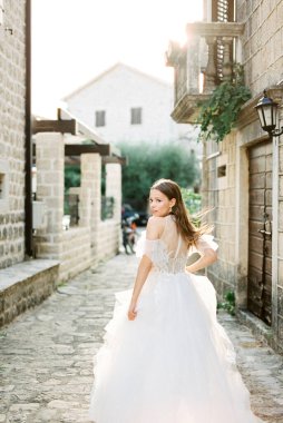 Bride stands looking back on a narrow cobbled street between ancient houses. Back view. High quality photo