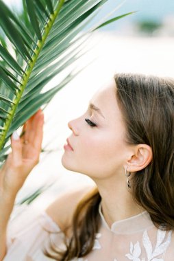 Bride in a white lace dress touches a green palm branch. High quality photo