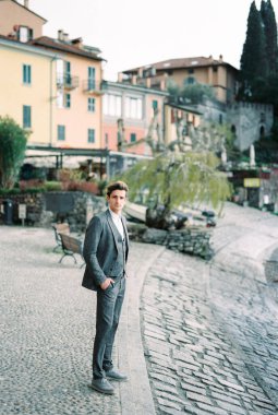 Man in a suit stands on a paved embankment with houses in the background. Varenna, Italy. High quality photo
