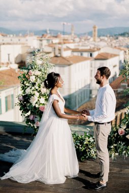 Bride and groom are standing near the wedding semi-arch of flowers on the terrace of the building. High quality photo