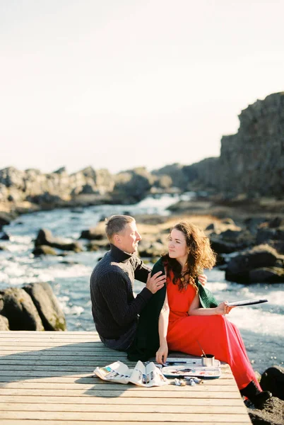 Man and a woman with an easel and paints are sitting on a wooden bridge near the river. High quality photo