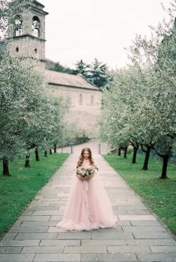 Bride with a bouquet of flowers walks along a paved path in an olive grove. High quality photo