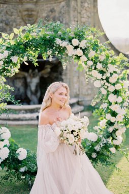 Bride in a veil with a bouquet stands at the wedding arch. High quality photo