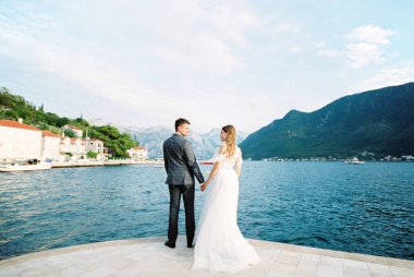 Bride and groom stand holding hands on the pier against the backdrop of Perast. Montenegro. High quality photo