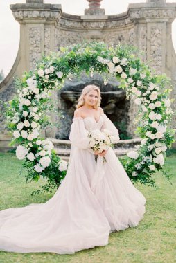 Bride with a bouquet stands at the wedding arch near the stone fence. High quality photo