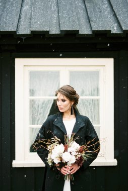 Bride in a jacket with a bouquet stands near a black wooden house. Iceland. High quality photo