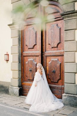 Bride in a white dress with a veil stands near the wooden door of the building. High quality photo