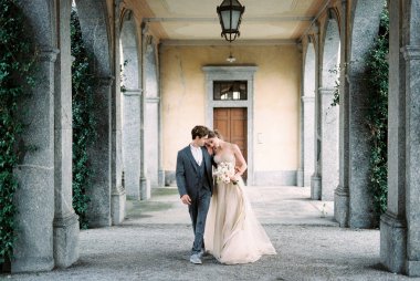Bride hugs groom, walking on the terrace with arches in the garden. High quality photo