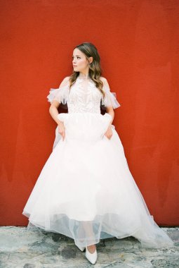 Bride in a white dress stands near a red wall. High quality photo