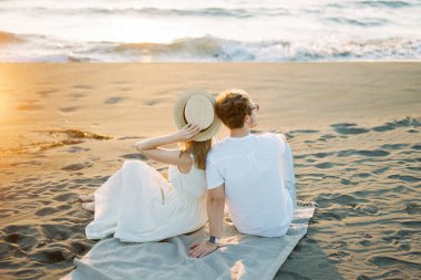 Woman in a hat sits on a blanket on the beach, leaning against a man shoulder. High quality photo