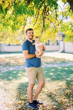 Dad with a baby in his arms stands under a tree in the park. High quality photo