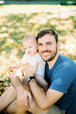 Smiling dad sitting with baby on lap in park. High quality photo