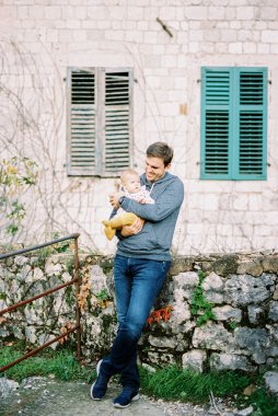 Dad with a baby in his arms stands at the stone wall of the house leaning on the foundation. High quality photo