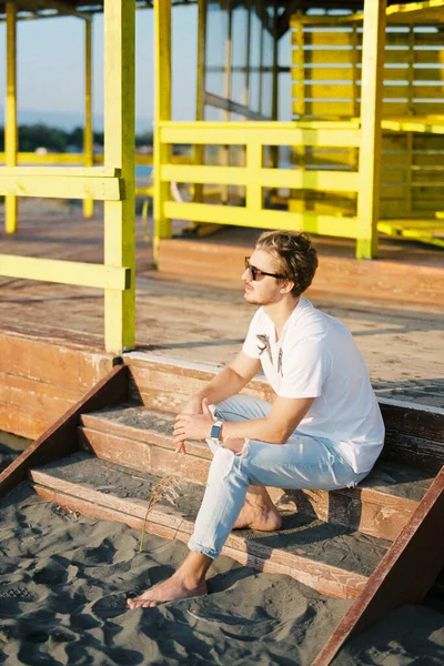 Young man in sunglasses sitting on the steps of a wooden building on the beach. High quality photo