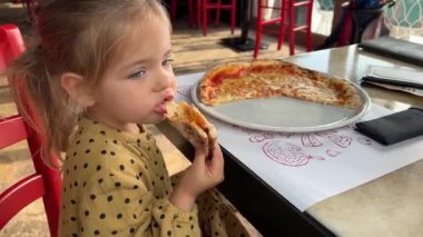 Little girl eating pizza holding a piece in her hand at a table in a cafe. High quality FullHD footage