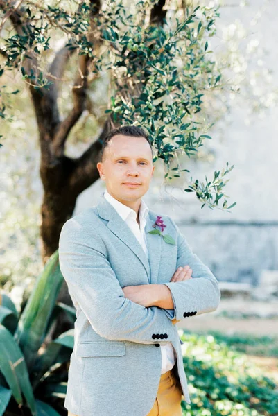 Groom in a jacket with a boutonniere stands with crossed arms under a tree. High quality photo