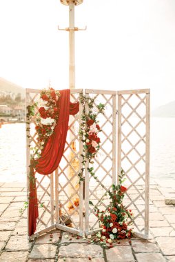 Kırmızı güller ve kumaşlarla süslenmiş ahşap düğün perdesi deniz kıyısındaki bir rıhtımda duruyor. Yüksek kalite fotoğraf
